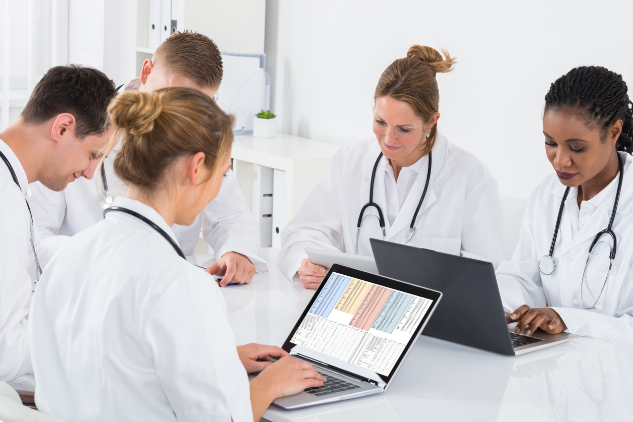 A clinical data analyst works on a laptop displaying a spreadsheet at a table with care providers.