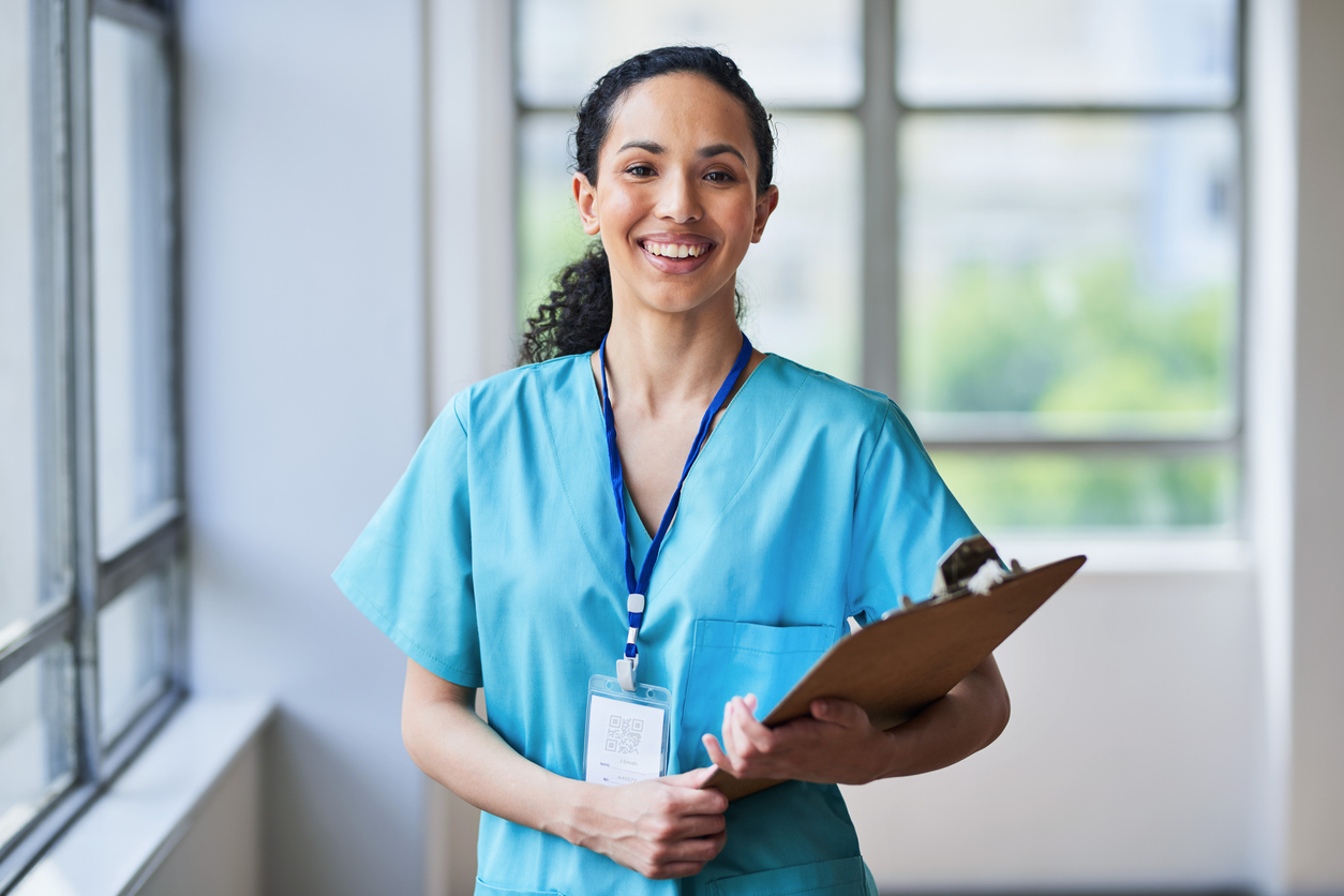 A smiling nurse holding a clipboard in a hospital corridor.