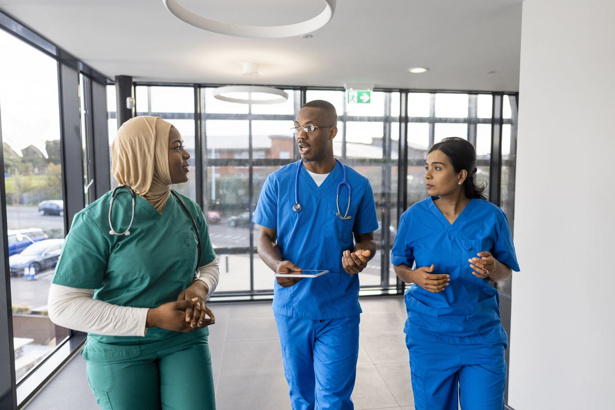 Three nurses in scrubs, one wearing a hijab, talk in a hospital corridor.
